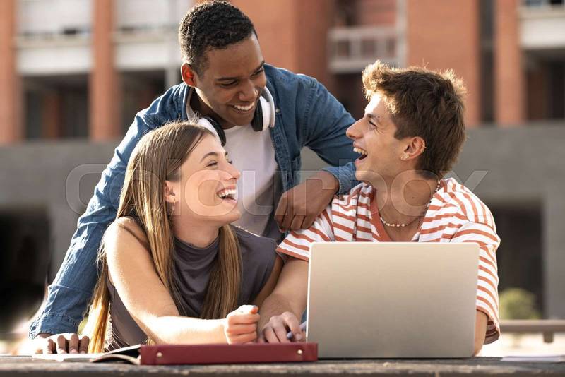 Cheerful group of people working together with a laptop in campus.Young multiracial friends studying and laughing outside using computer and notes.
