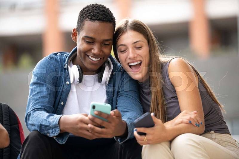Happy young couple laughing sitting on stairs using their phones.Two cheerful diverse friends laughing and texting outside.