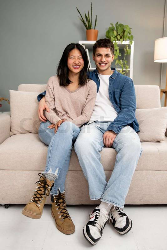 Lovely affectionate smiling young adult couple sitting on sofa at home looking at camera. Diverse carefree man and woman hugging and relaxing together in couch at the living room.