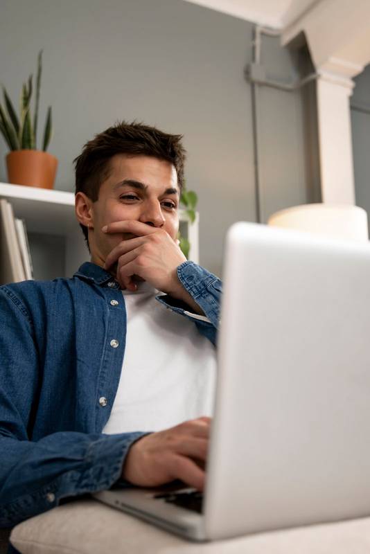 Excited young adult man laughing while using a laptop at home. Surprised guy working with a computer in his living room.