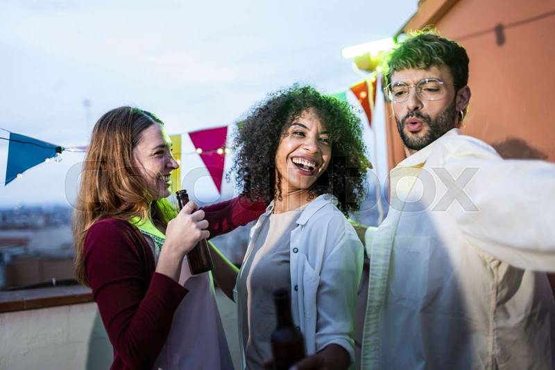 Three friends dancing together in a rooftop party laughing at evening. Young happy millennials having fun in a terrace and having drinks