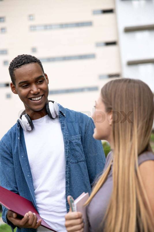 Multiracial couple smiling and holding folders in a campus.Two young students looking each other laughing and relaxed in a park.