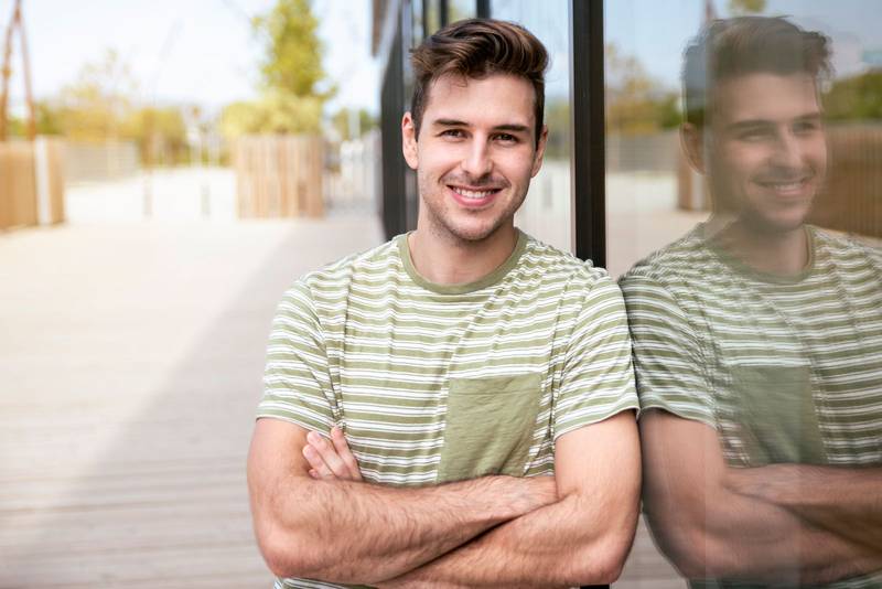 Portrait of young man smiling happy with arms crossed at the city standing next to the glass wall