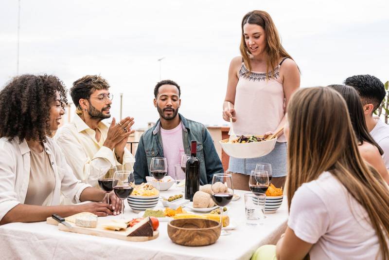 Young woman serving salad to her friends in a outdoors dinner party. Group of multiracial friends having rooftop dinner party together.