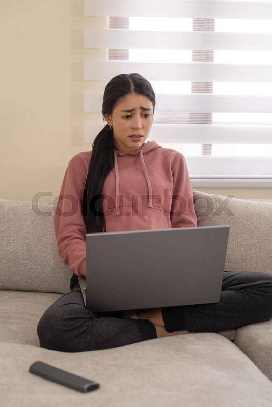 woman sits on a sofa with a concerned expression while using her laptop. Represents stress, bad news, or emotional discomfort