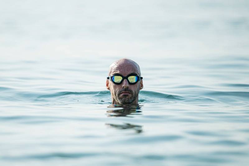 Confident man swimming in the ocean wearing glasses looking at camera. Swimmer pulling his head out of the water focused.