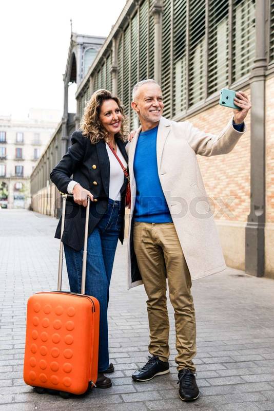 Happy mid adult tourist couple travelling together taking a selfie with her luggage. Affectionate senior husband and wife taking a picture during a weekend romantic trip in european city