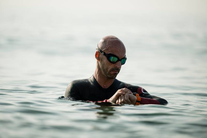 Confident man swimming in the ocean wearing glasses looking at his watch. Swimmer standing in the water checking his time with a smartwatch.