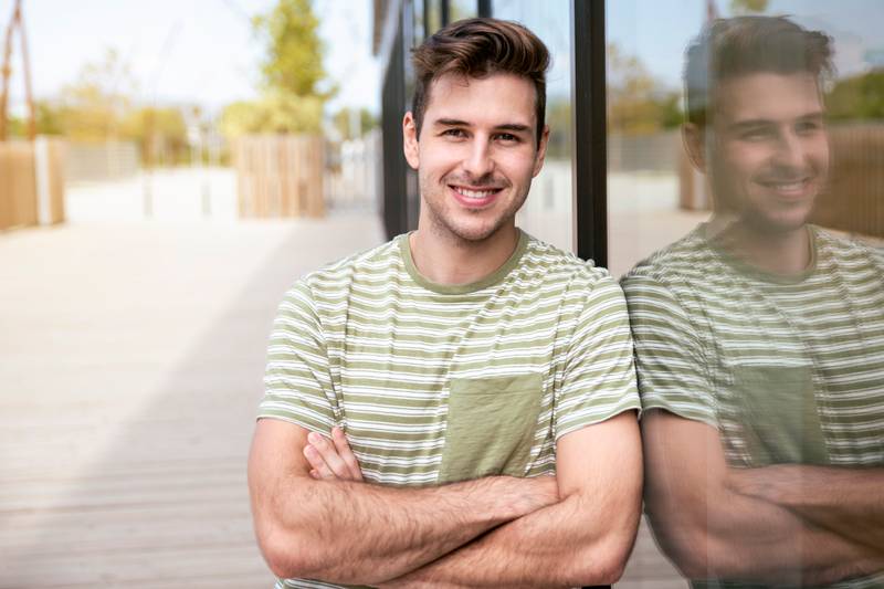 Portrait of young man smiling happy with arms crossed at the city standing next to the glass wall