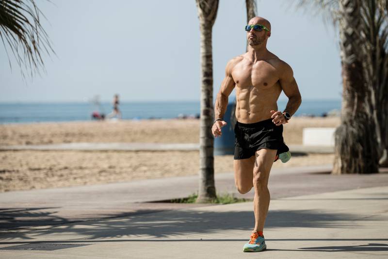 Athletic handsome man running in the street wearing sunglasses. Fit athlete jogging outdoors shirtless next to the beach
