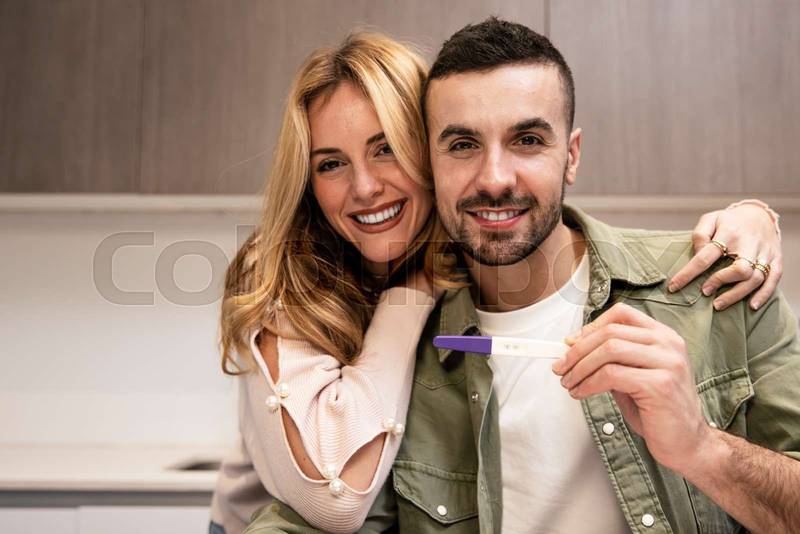 Affectionate happy young couple showing positive pregnancy test and hugging at home. Handsome young adult man holding maternity test with his wife embracing looking at camera.