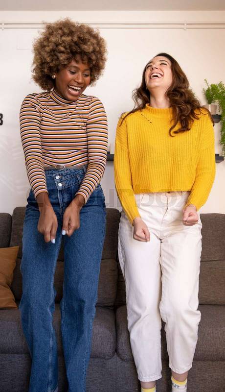 Multiracial young funny women dancing and laughing in their apartment. Happy and excited couple of girls celebrating in their living room.