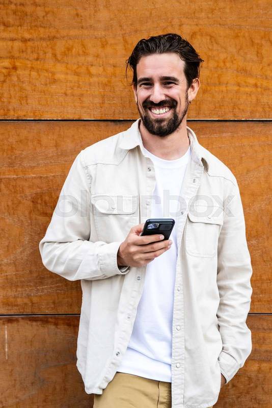 Confident handsome young guy texting with his phone and looking at camera. Happy and relaxed man using his smartphone standing on a brown wall outside staring at camera.