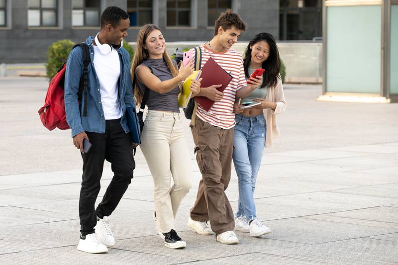 Young group of students walking and using phones in the street. Diverse students texting and laughing while moving in the campus.