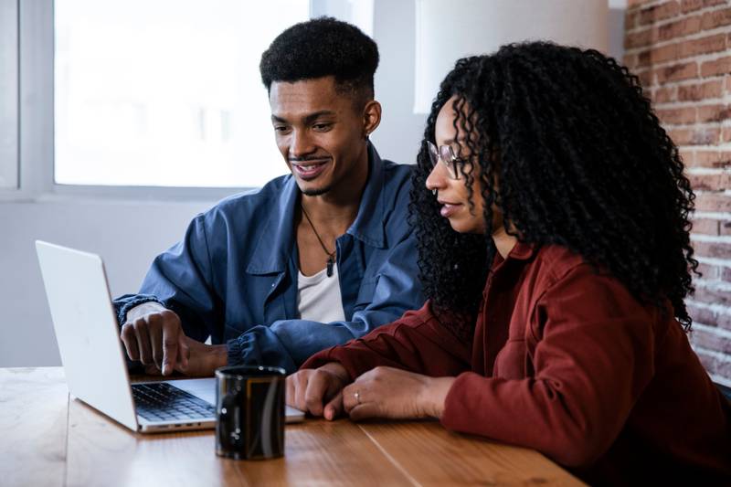 Handsome young couple using laptop together while sitting at home. Smiling friends looking at computer and laughing at cozy home office.