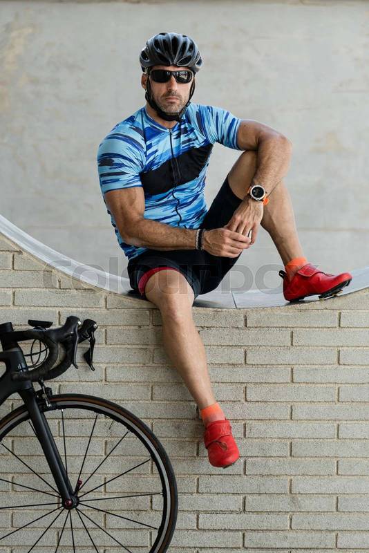 Bike rider in sport clothes sitting in a wall with a cycle looking at camera. Fit cyclist resting next to his cycle wearing helmet and sunglasses.