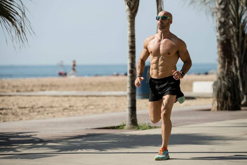 Athletic handsome man running in the street wearing sunglasses. Fit athlete jogging outdoors shirtless next to the beach