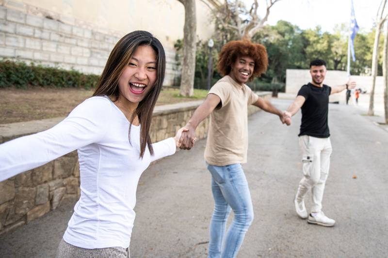 Three diverse and confident friends holding hands together. Multiracial group of people laughing and holding hands in a line. Focus on first woman.