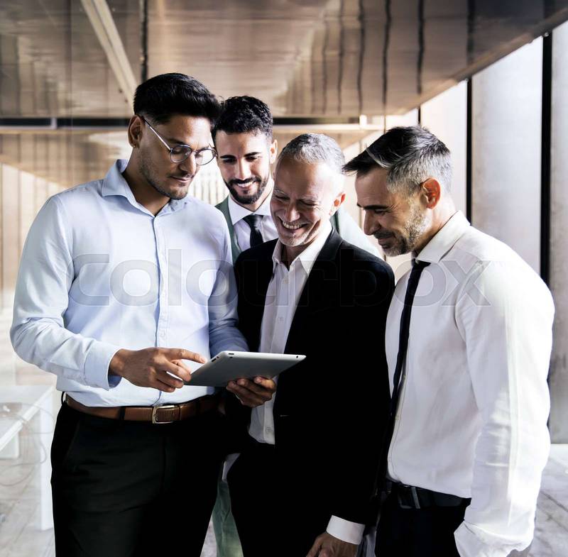 Diverse group of businessmen checking a tablet in an office corridor. Young executive holding a device in a meeting with senior colleagues standing at office.