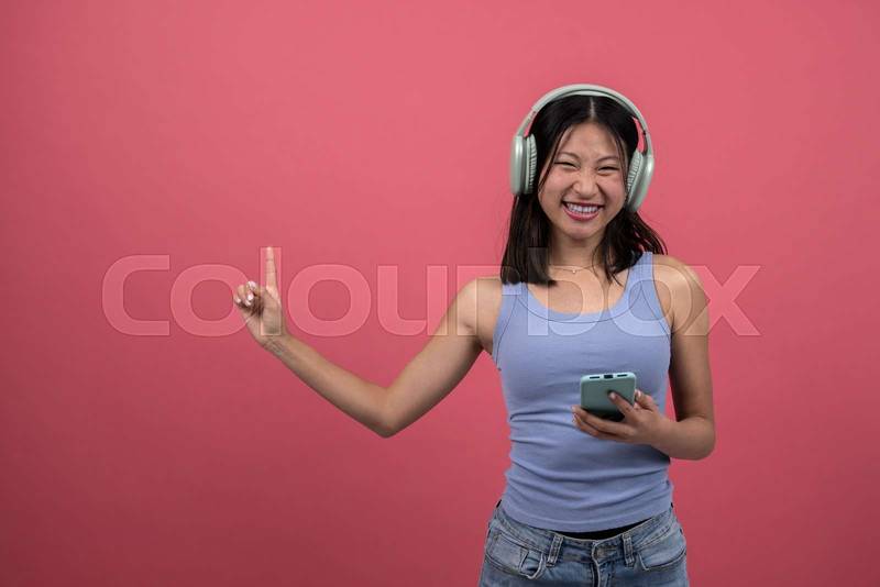Joyful woman listening radio with phone pointing up. Young happy female using headphones listen to music with smartphone isolated on red background studio portrait.