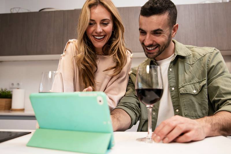 Happy joyful man and woman on a video call having a glass of wine. Young adult couple using together a tablet from home in a modern kitchen.