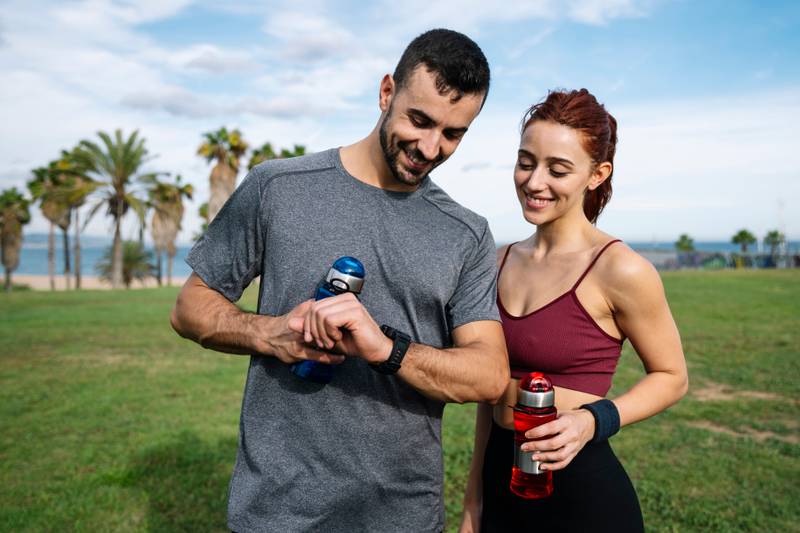 Runners resting after training looking at sports watch checking heart rate. Joyful fit couple jogging in park checking smartwatch while holding water bottles outside