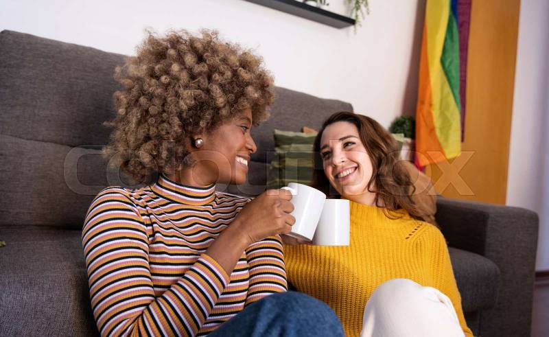 Multiethnic couple taking a coffee break sitting in the floor of a living room. Two cheerful women looking each other while toasting with cups in their apartment.