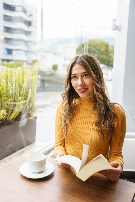 sitting relaxed a latin young woman with long blonde hair, enjoys a cup of hot drink and reads