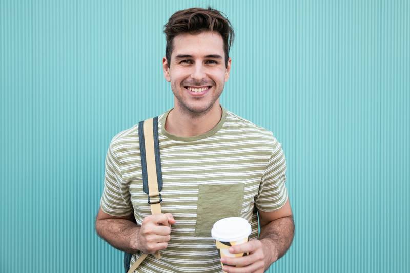 Portrait of handsome happy student smiling and drinking a take away coffee at a green wall.