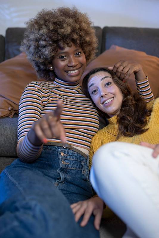 Multiracial cheerful lesbian couple hugging and pointing to camera. Close up of smiling young gay females looking at camera while relaxing in a living room.