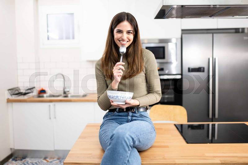 Portrait of beautiful young woman having breakfast in the kitchen. smiling female with a bowl ans spoon looking at camera. Lifestyle concept.