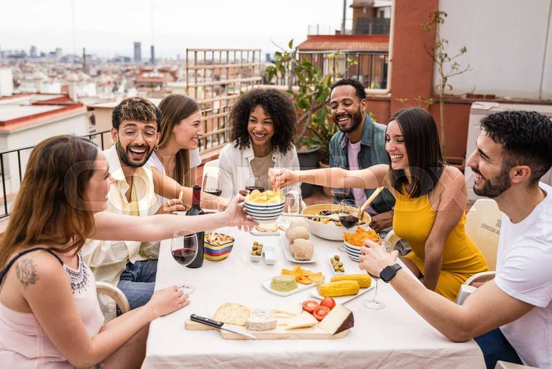 Cheerful group of diverse friends eating and drinking at a terrace table. Young and relaxed people having fun in a rooftop dinner party.