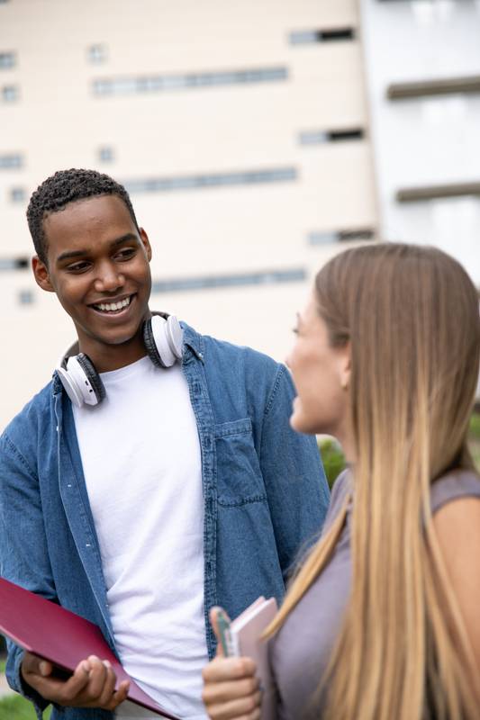 Multiracial couple smiling and holding folders in a campus.Two young students looking each other laughing and relaxed in a park.