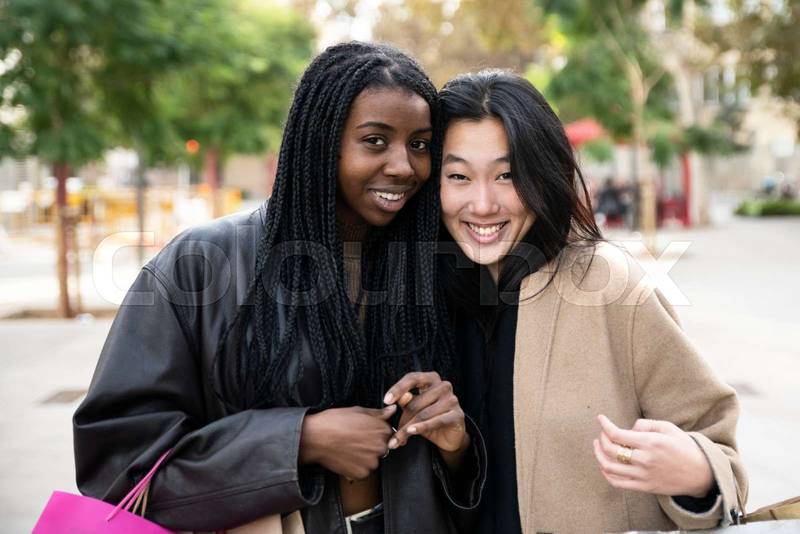Portrait of two beautiful young women looking at camera and holding shopping bags. Smiling multiethnic friends together in the street