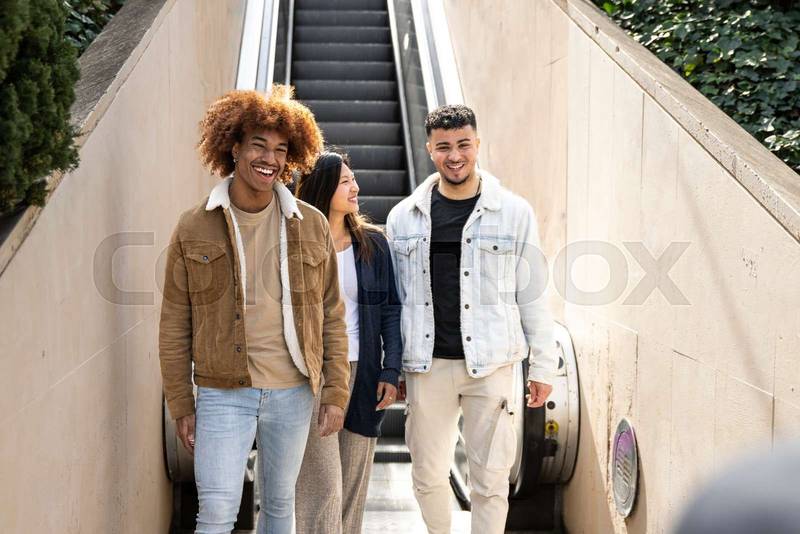 Diverse group of friends going down an outdoor escalator. Three happy young people smiling and looking relaxed walking down of stairs in the street.