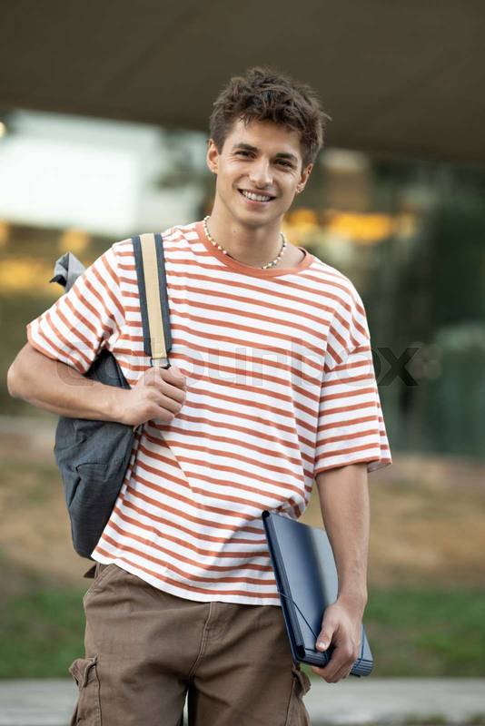 Handsome student wearing a bacpack and holding a folder looking at camera.Relaxed and happy young man standing outside with files.