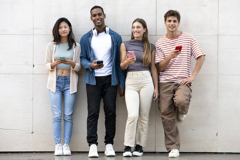 Diverse group of young people texting leaning against a wall looking at camera. Happy group of millennials friends using their phones standing in the street staring at the camera.