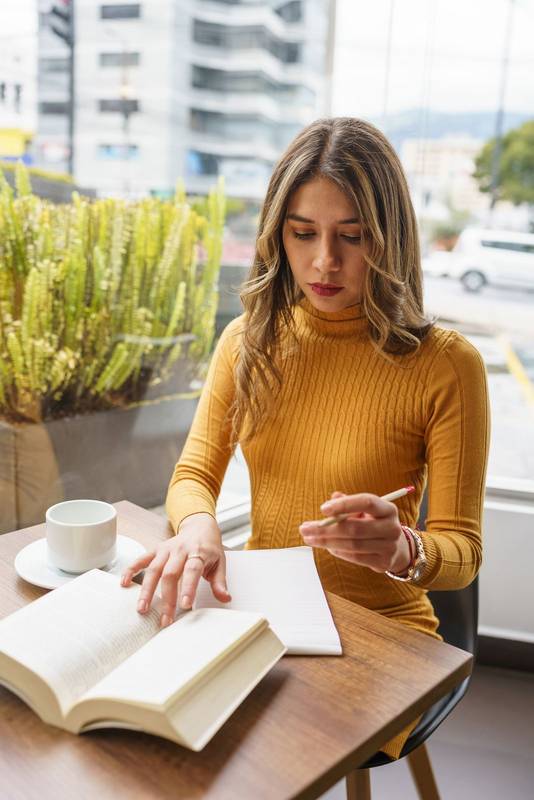 day of young female student with blonde hair, learning with a book and taking notes with a cup of hot drink
