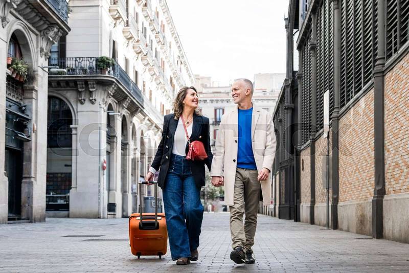 Affectionate mid adult tourist couple walking together with luggage in a european city. Lovely happy senior man and woman traveling with a suitcase visiting streets of old town.