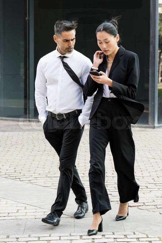 Businesswoman and businessman checking a phone and walking in the street. Casual meeting between a young executive and a senior ceo while texting on the smartphone.