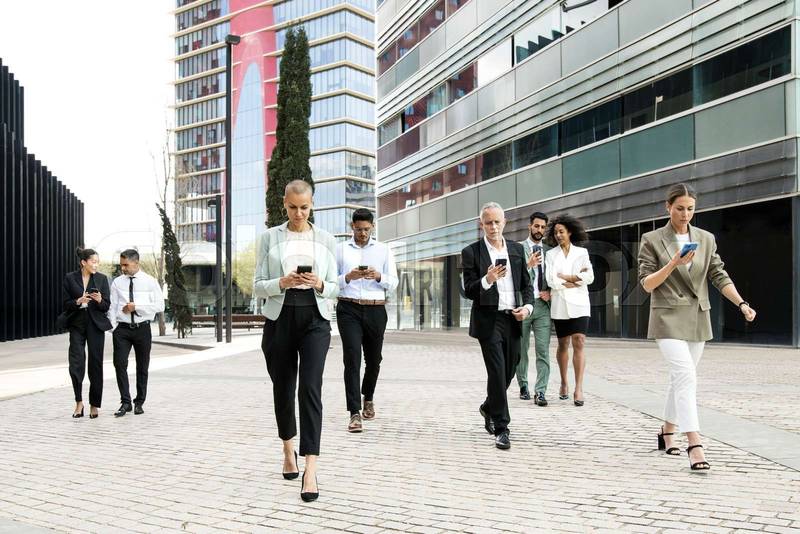 Group of confident business people walking the street using their phones. Diverse team of focused businesswoman and businessman walking outside while texting on their smartphones.