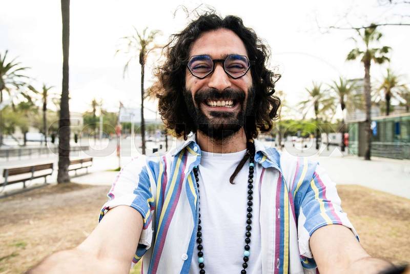 Portrait of happy hipster guy smiling wearing a shirt looking at camera and taking a selfie in the street. Carefree cheerful stylish male wearing glasses confident and satisfied standing outside 