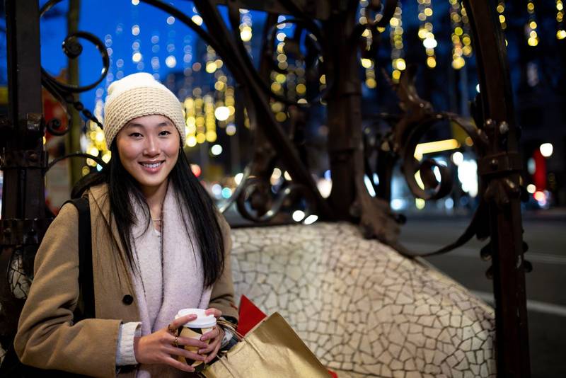 Joyful young woman shopping at evening looking at camera. Cheerful female holding a coffee standing at night in the street in winter.