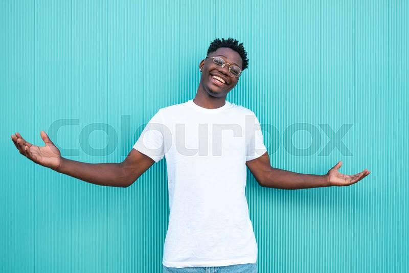 Portrait of a happy african american guy smiling with his arms wide open on turquoise background and white t-shirt.