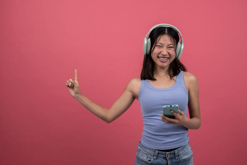 Joyful woman listening radio with phone pointing up. Young happy female using headphones listen to music with smartphone isolated on red background studio portrait.