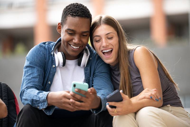 Happy young couple laughing sitting on stairs using their phones.Two cheerful diverse friends laughing and texting outside.