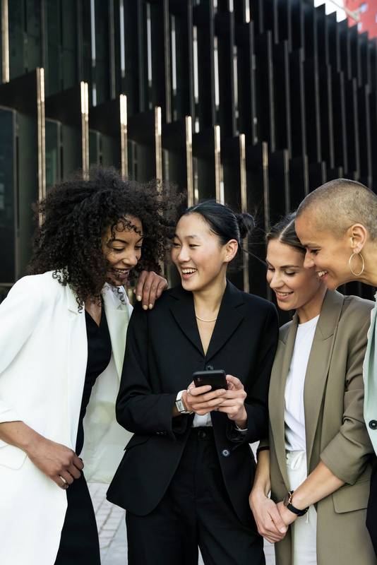 Diverse businesswomen group looking a phone and laughing standing in the street. Casual meeting between young females executives checking their smartphone outisde.