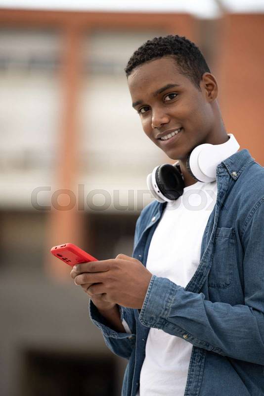 Young handsome guy smiling and looking at camera relaxed using his phone.Relaxed millennial man smiling and texting wearing headphones.