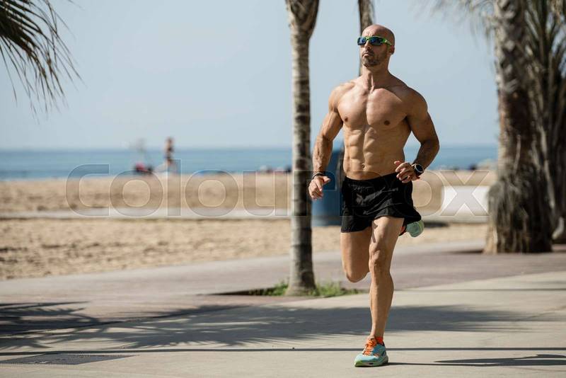 Athletic handsome man running in the street wearing sunglasses. Fit athlete jogging outdoors shirtless next to the beach