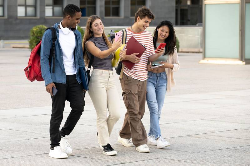 Young group of students walking and using phones in the street. Diverse students texting and laughing while moving in the campus.